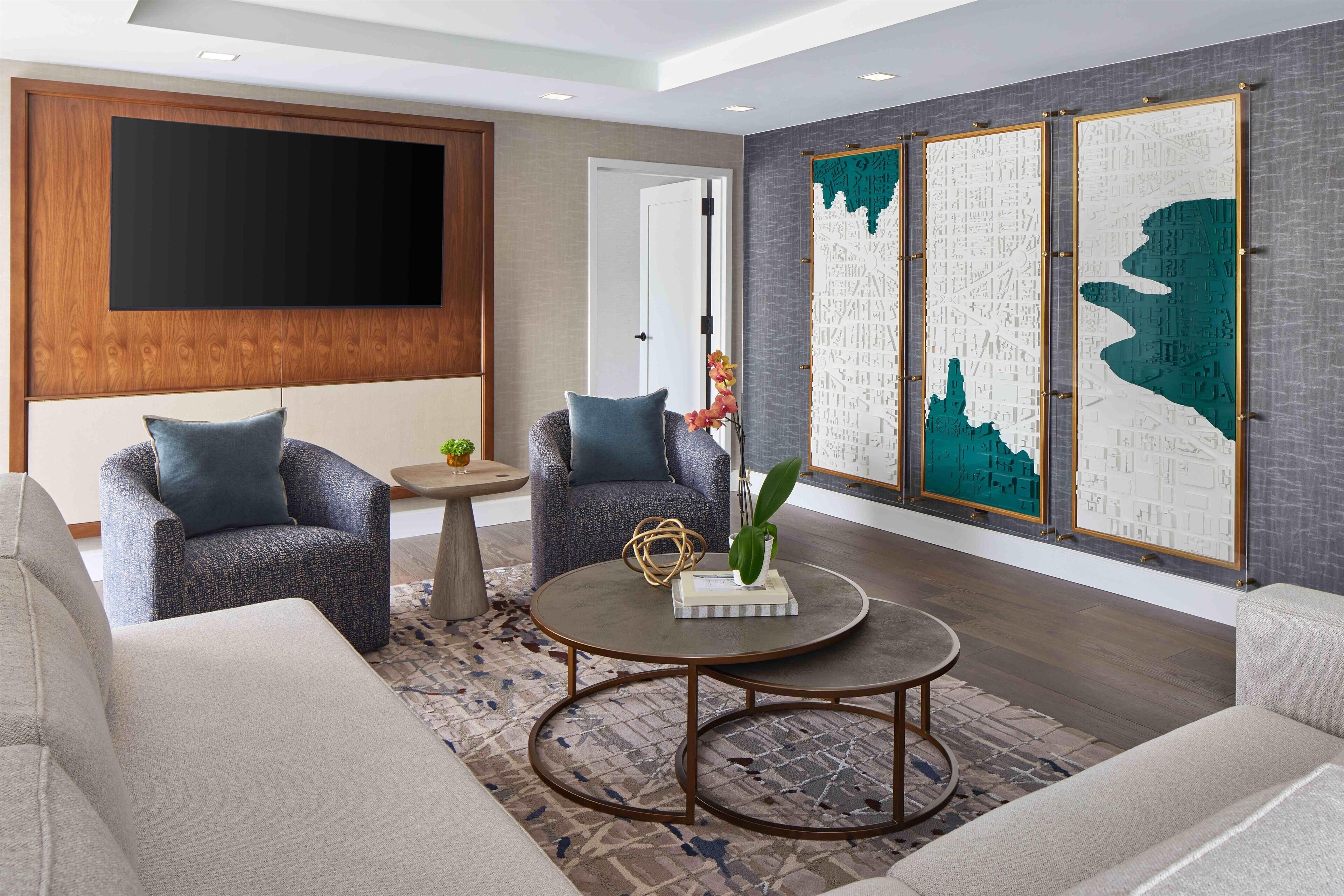 A modern living room with two grey armchairs, a round coffee table, and a large flat-screen TV mounted on a wooden panel wall.
