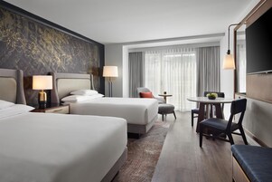 A hotel room with two beds, a dining table, a chair, a TV, and a large window with curtains.