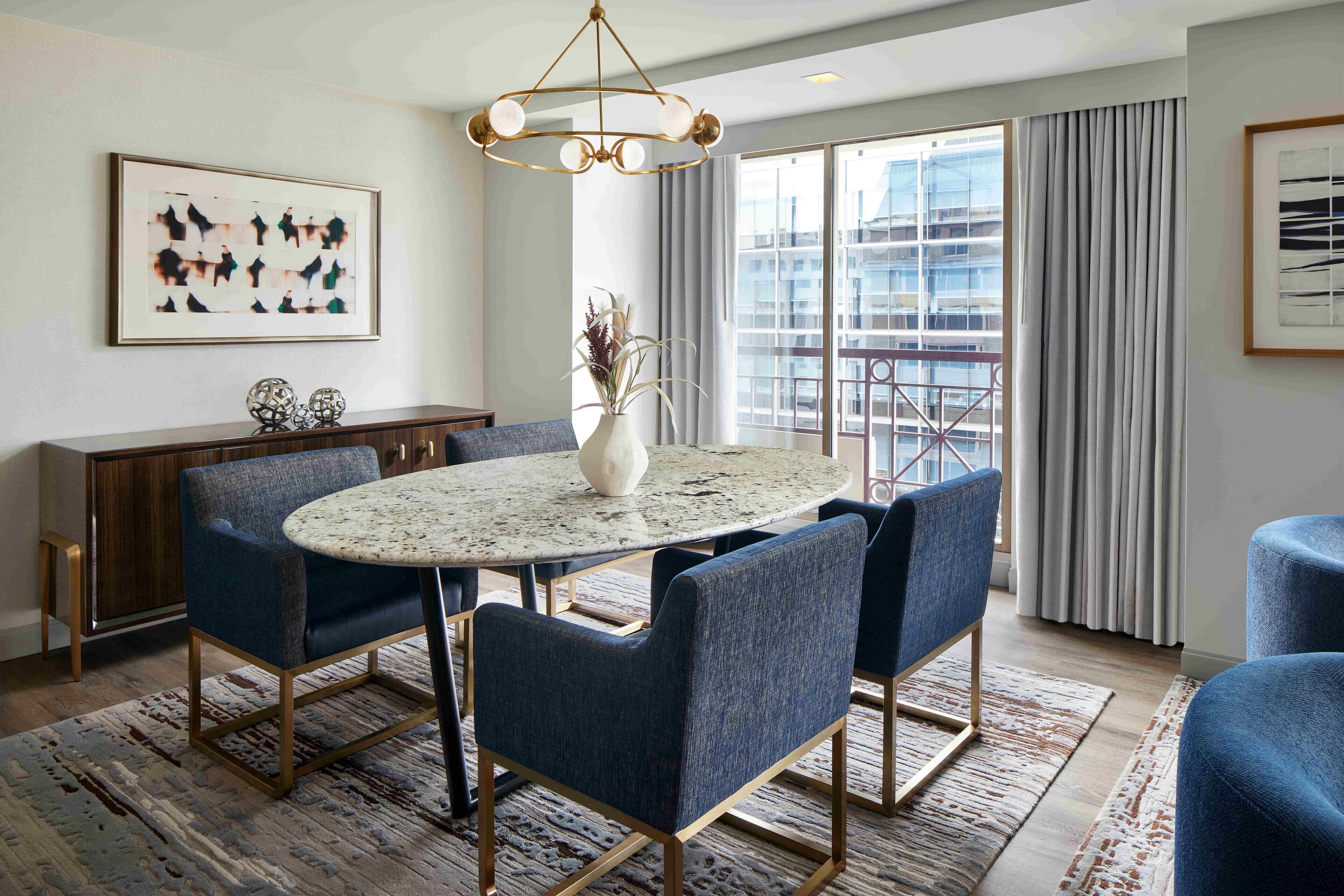 Uma sala de jantar com mesa redonda, cadeiras estofadas em azul, lustre dourado e vista para a paisagem urbana através de grandes janelas.