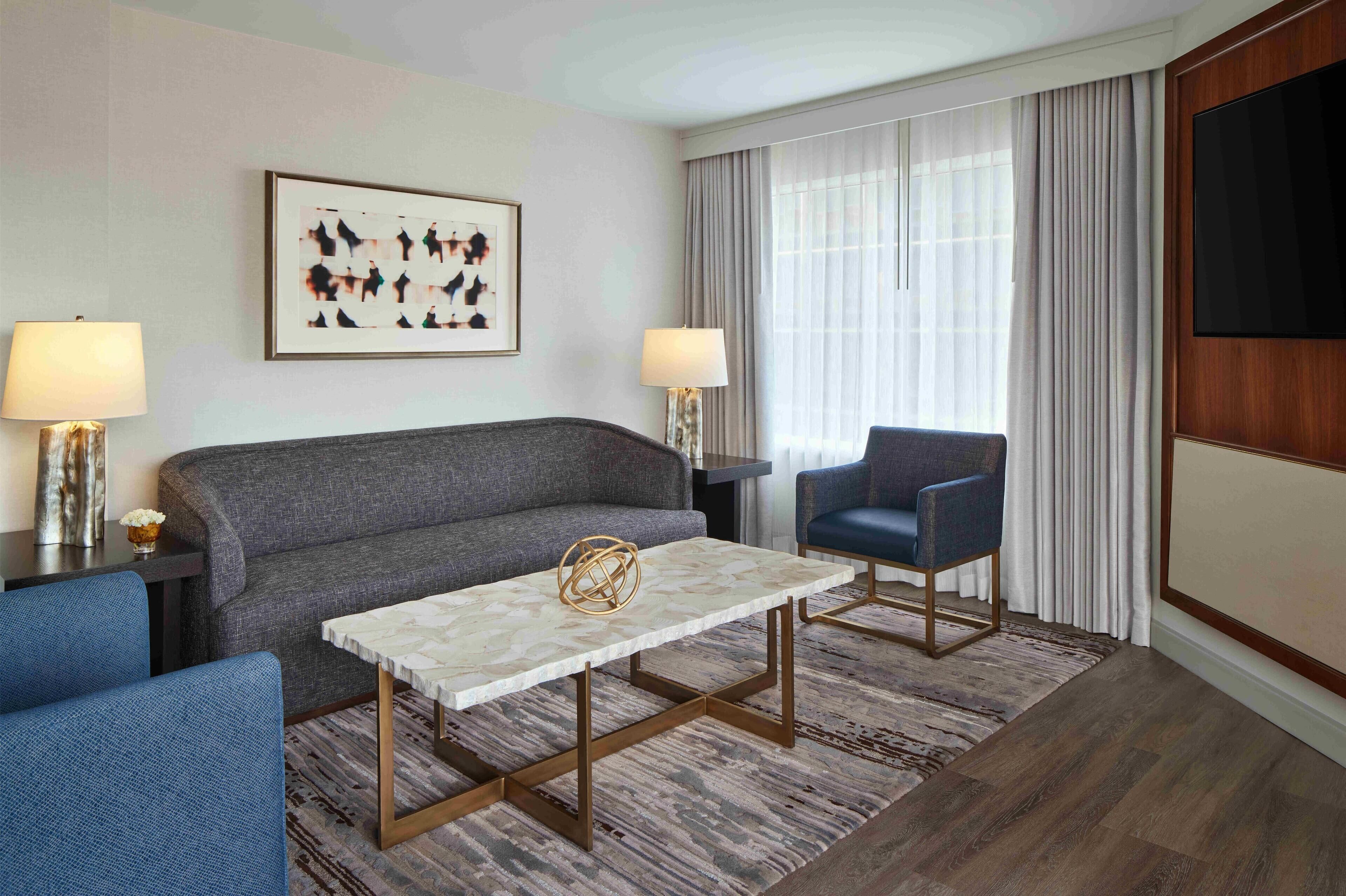 A modern hotel room with a grey sofa, a small table with a decorative object, two armchairs, a TV mounted on the wall, and a large window with curtains.