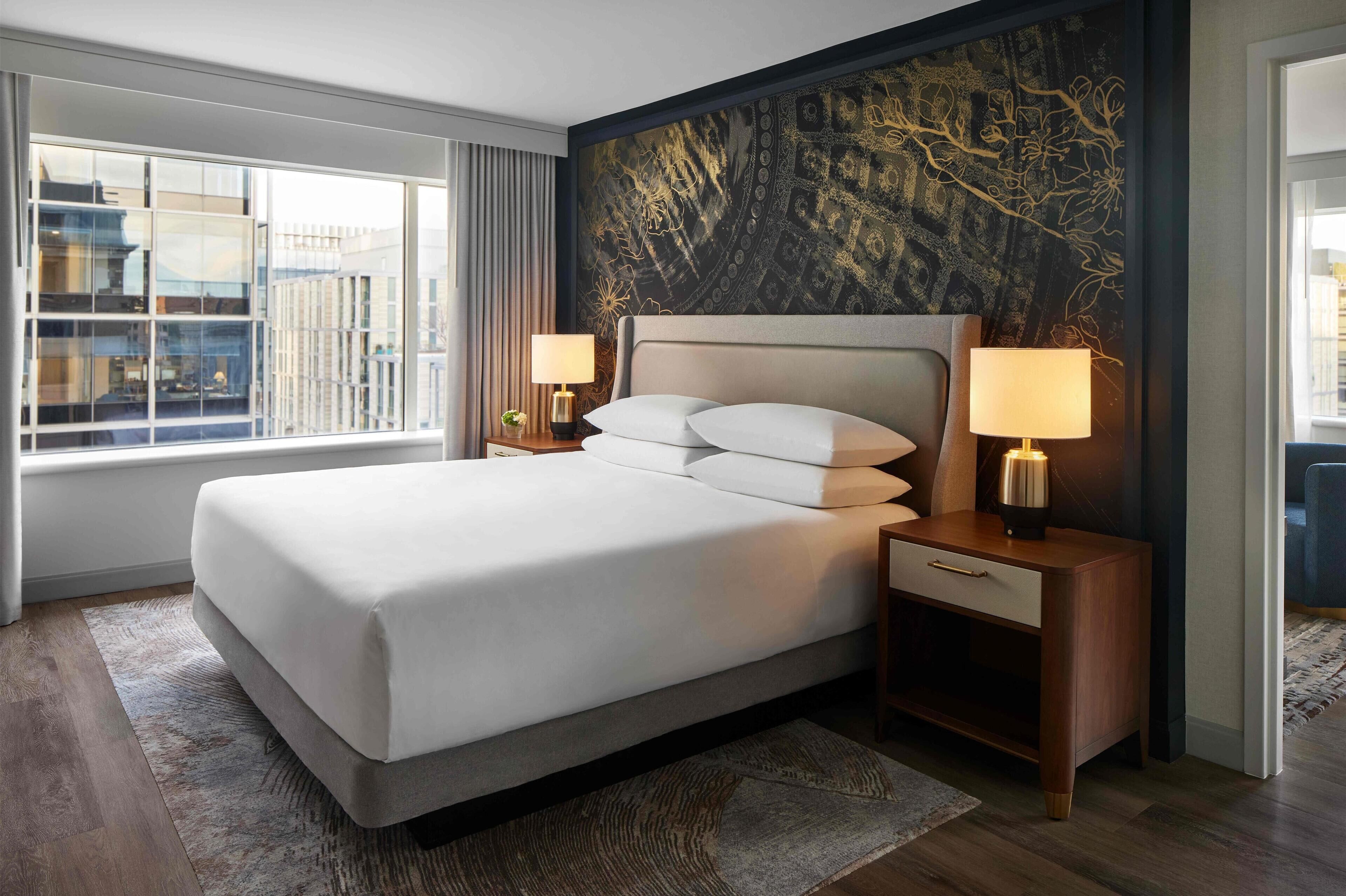 A modern hotel room with a large bed, two bedside tables with lamps, a view of the city through floor-to-ceiling windows, and a decorative wall mural.