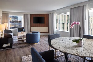 A modern hotel room with a large window, a flat-screen TV mounted on the wall, a round dining table with a plant, and a comfortable seating area with a sofa and chairs.
