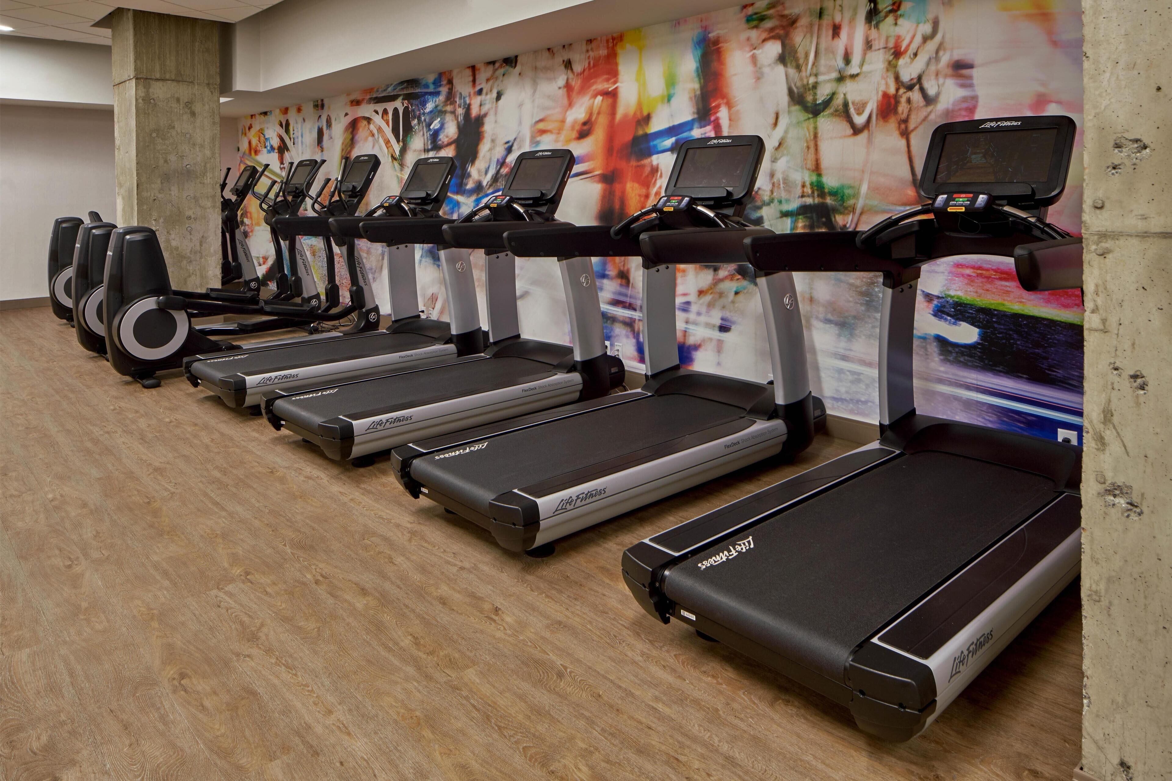 A row of treadmills in a gym with a colorful abstract mural on the wall.