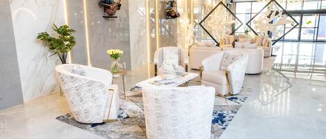 A modern lobby with marble walls, a seating area with white upholstered chairs, and a large window providing natural light.