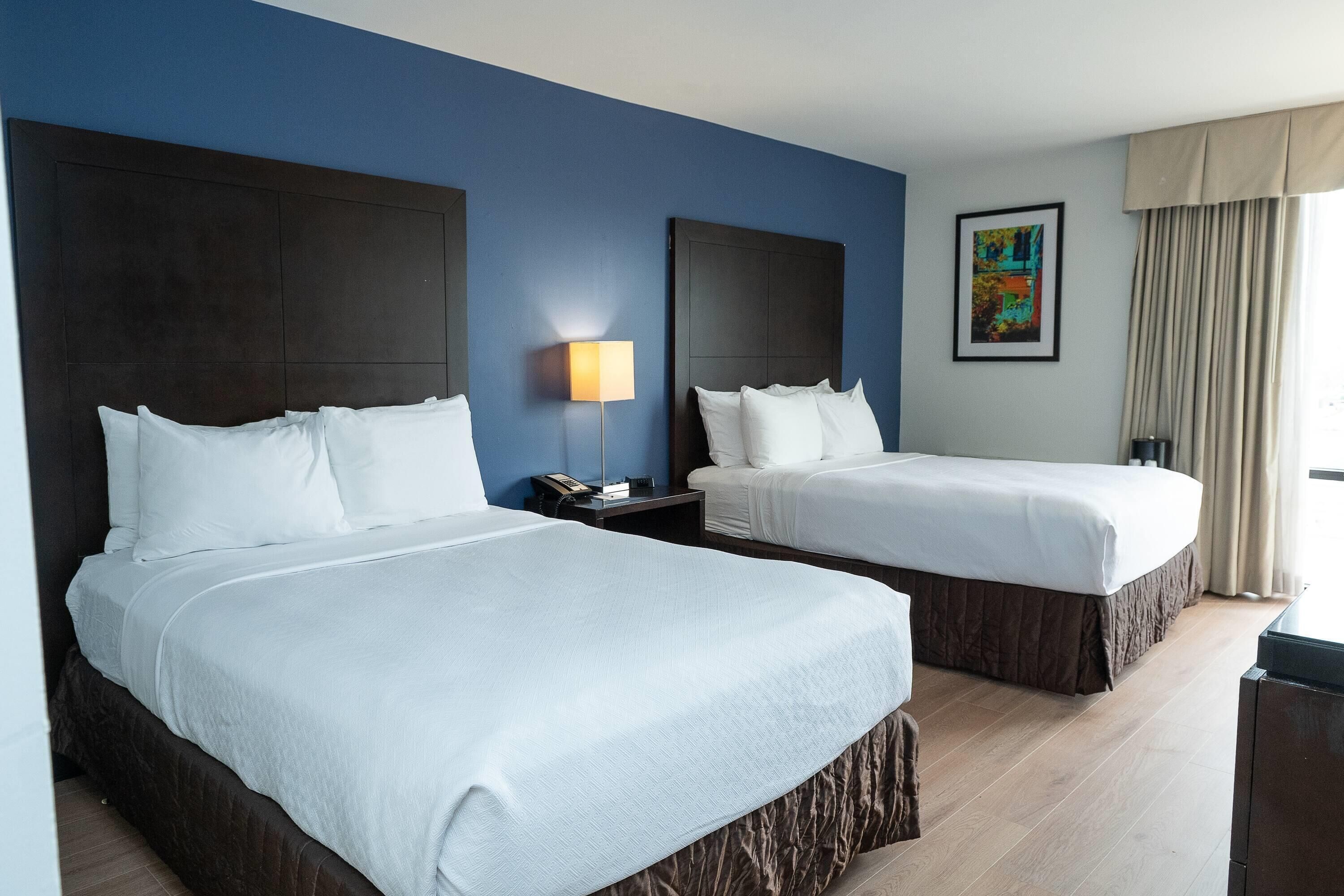 Habitación de hotel con dos camas, una mesita de noche con lámpara, un cuadro enmarcado en la pared y una ventana con cortinas.