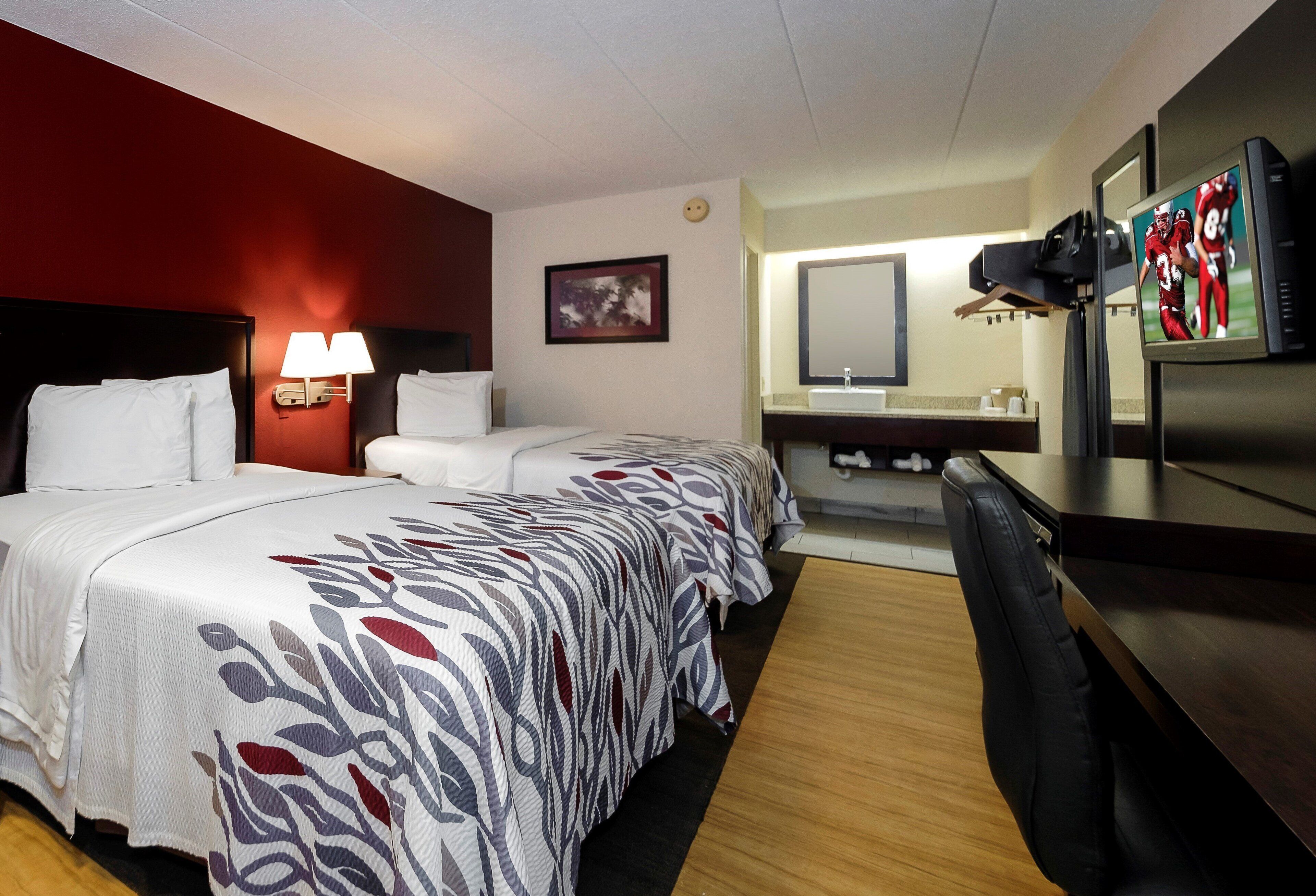 A hotel room with two beds, a desk, a chair, a TV, and a bathroom with a mirror and sink.