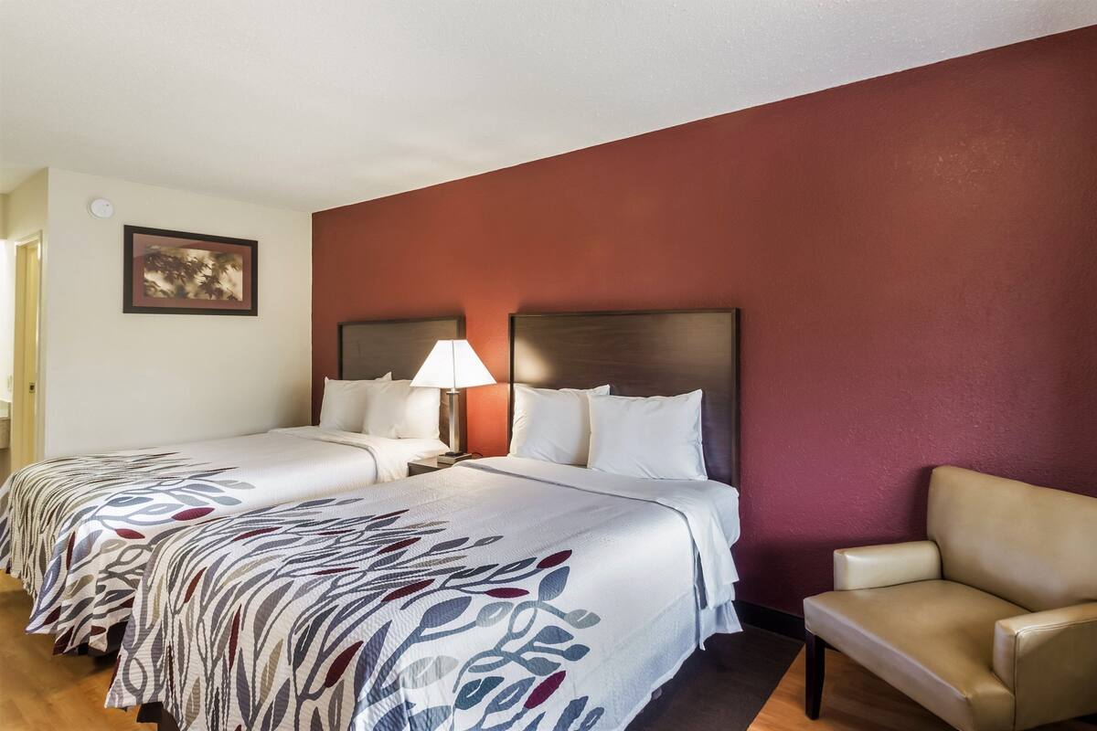 A hotel room with two beds, a nightstand with a lamp, a chair, and a painting on the wall.