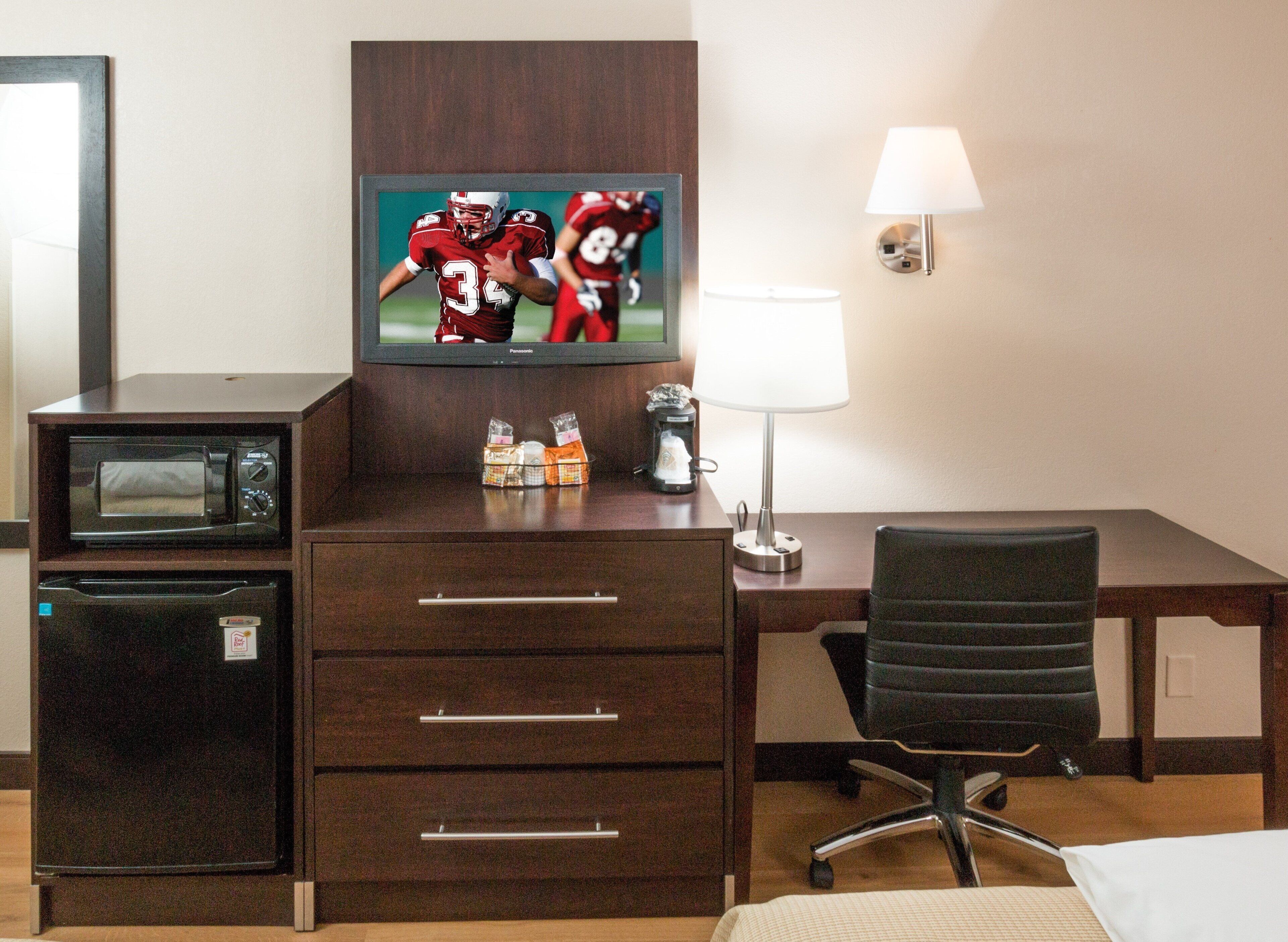 A hotel room with a television, a microwave, a refrigerator, a desk, a chair, and a bedside table.
