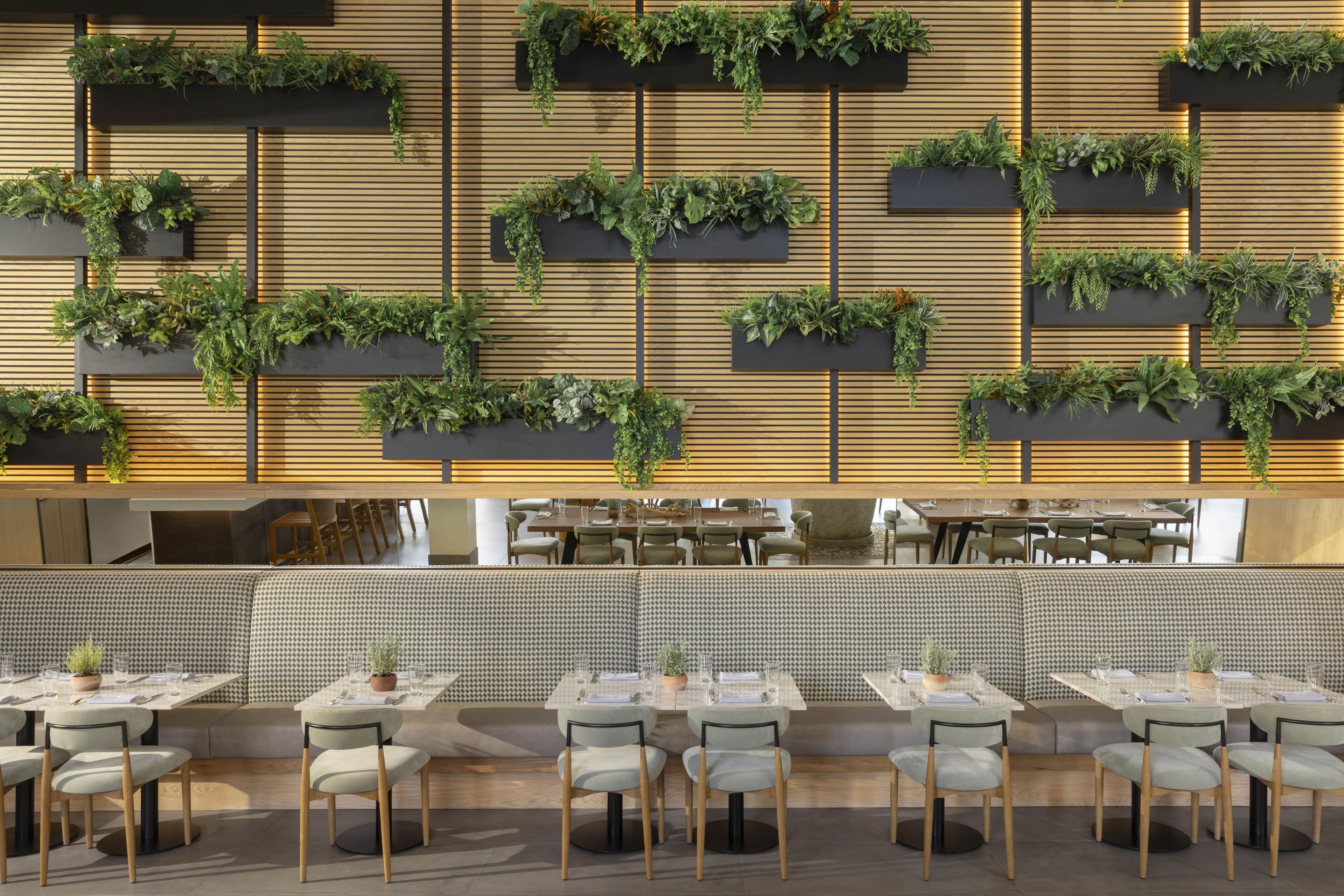 A restaurant interior with multiple dining tables and chairs, a long bench seating area, and vertical garden installations on the walls.