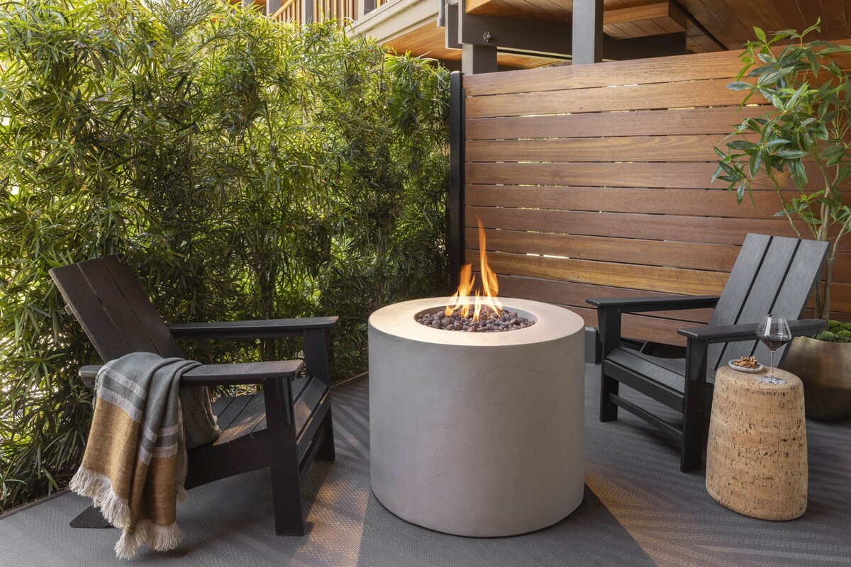 A circular fire pit with flames, surrounded by two chairs and a bench, set against a backdrop of greenery and a wooden fence.