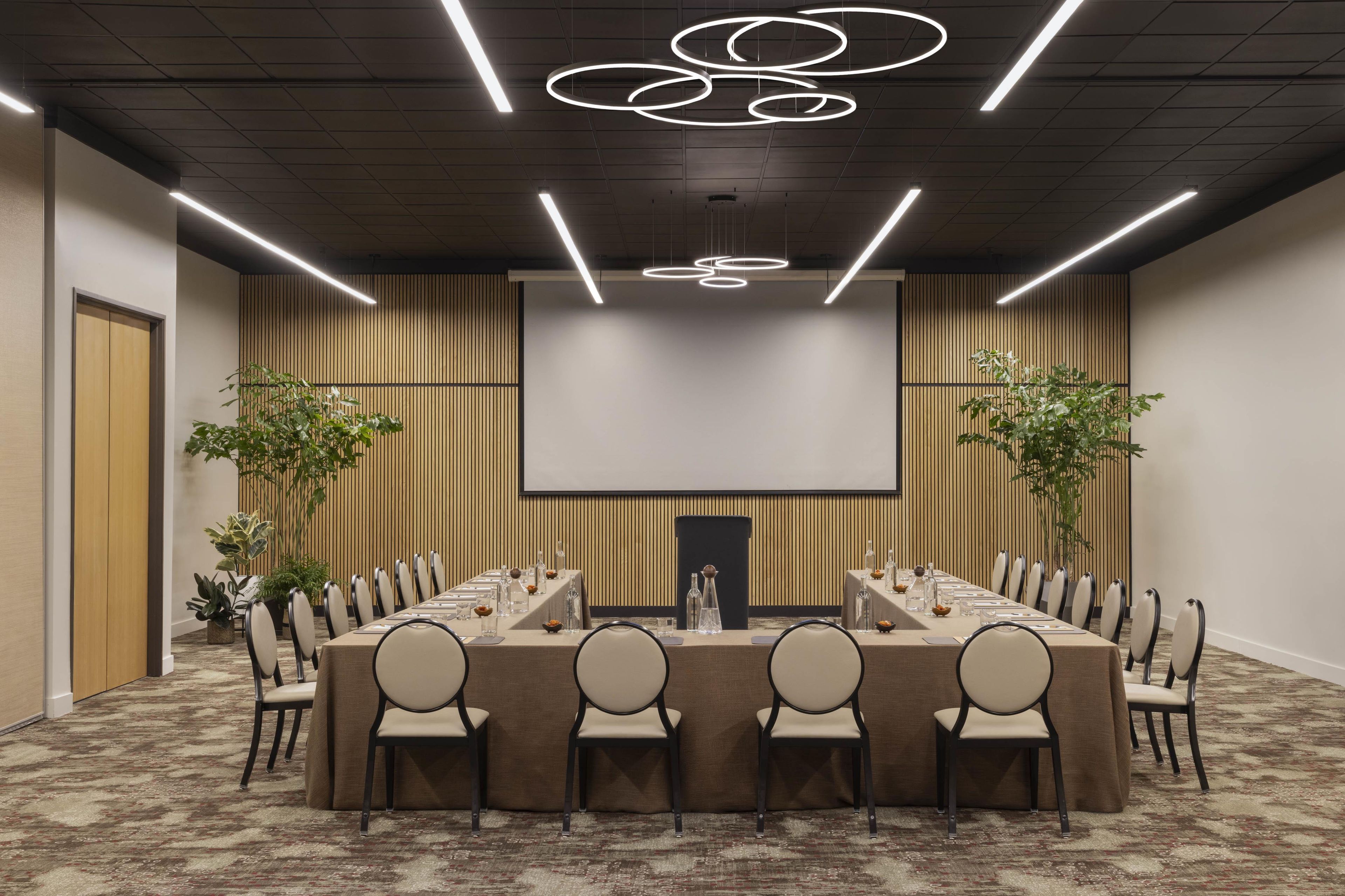 Un salón de conferencias con una larga mesa, sillas, una pantalla de proyector y un moderno candelabro.