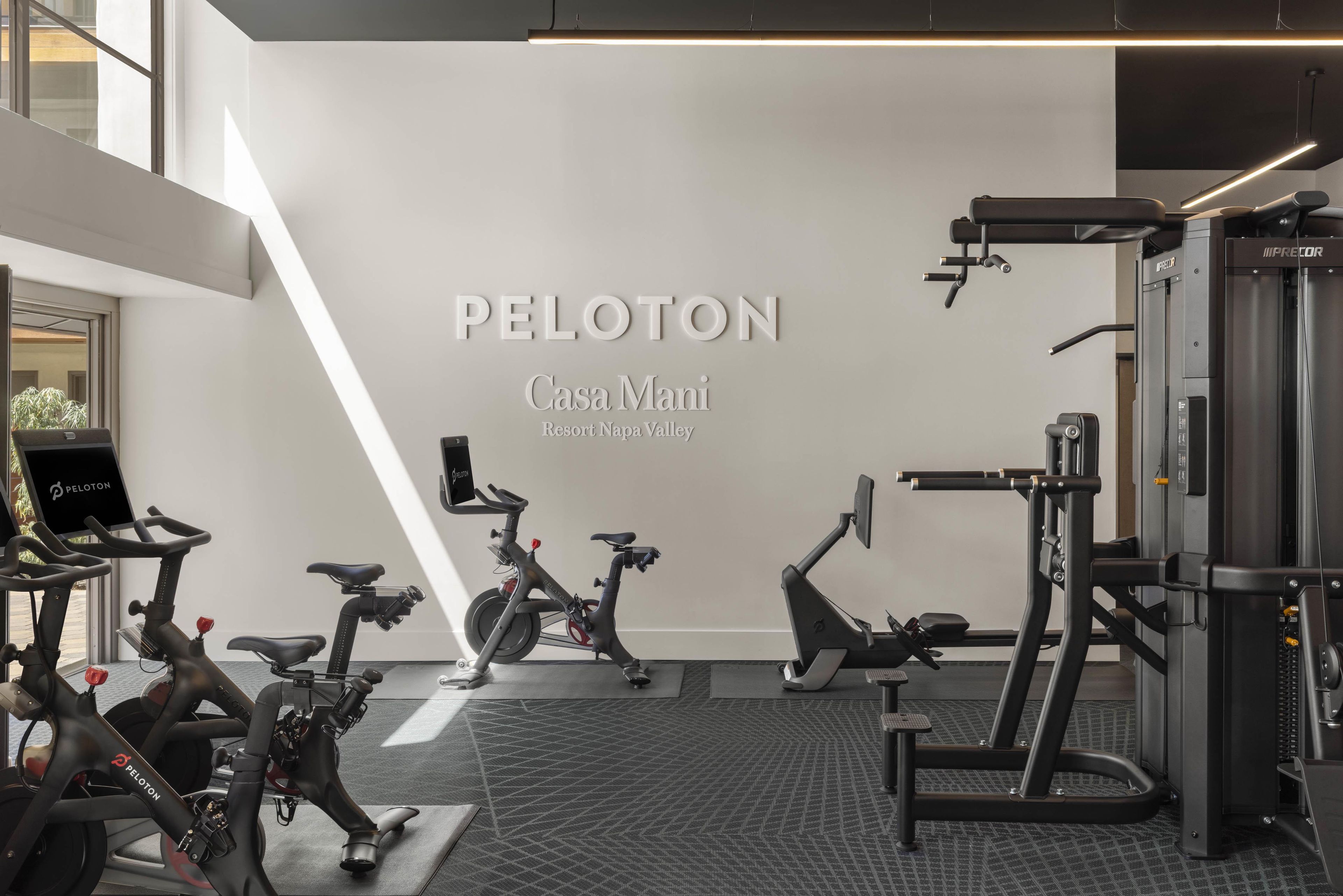 A Peloton exercise bike and a stationary bike in a gym with a wall-mounted Peloton logo and text.