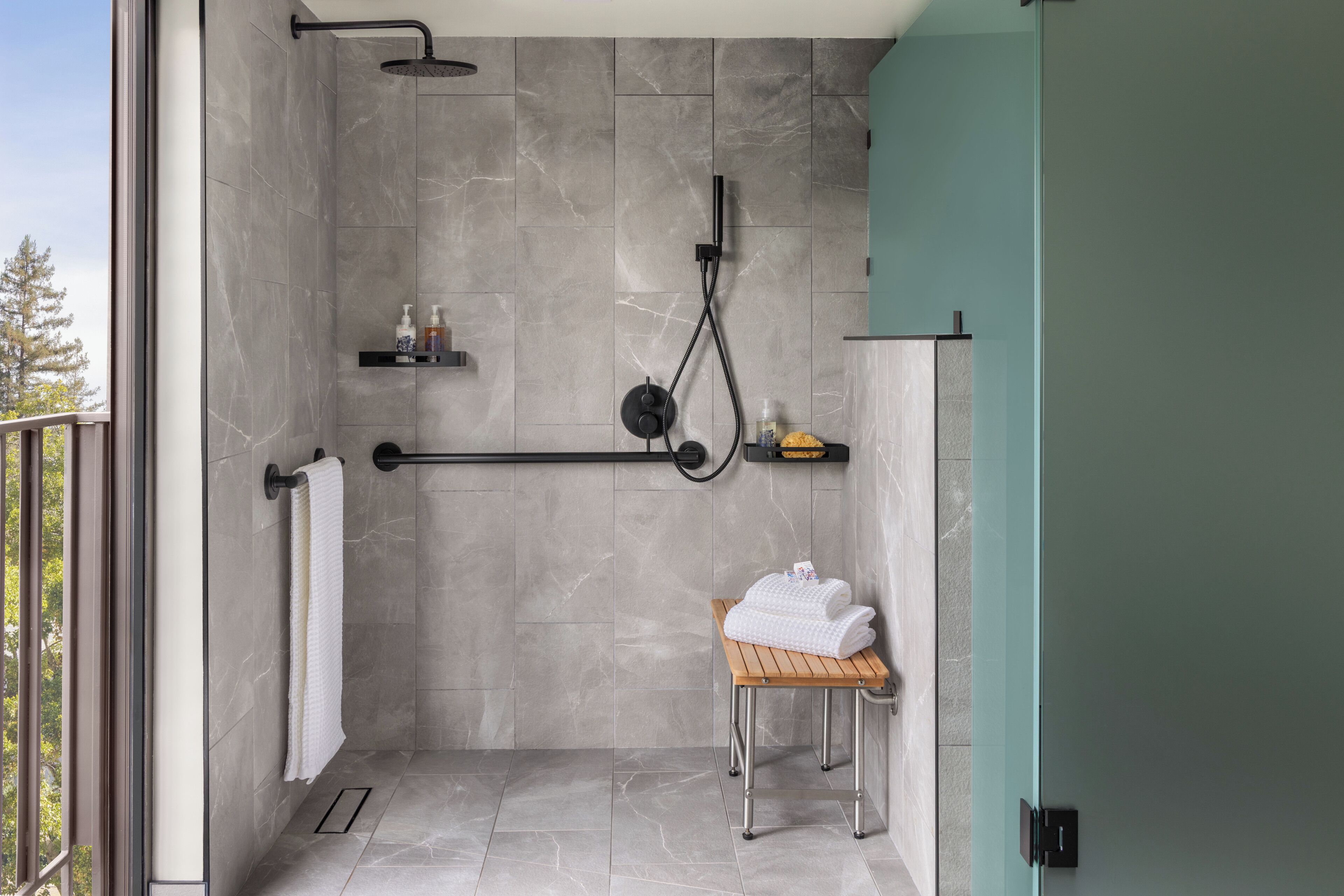 A modern shower area with a rain showerhead, a handheld showerhead, and a handheld massager. There is a wooden stool with towels and a soap dispenser on a shelf.
