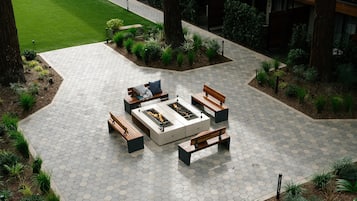 Un patio hexagonal con baldosas, con una chimenea central, rodeado de bancos de madera y macetas.