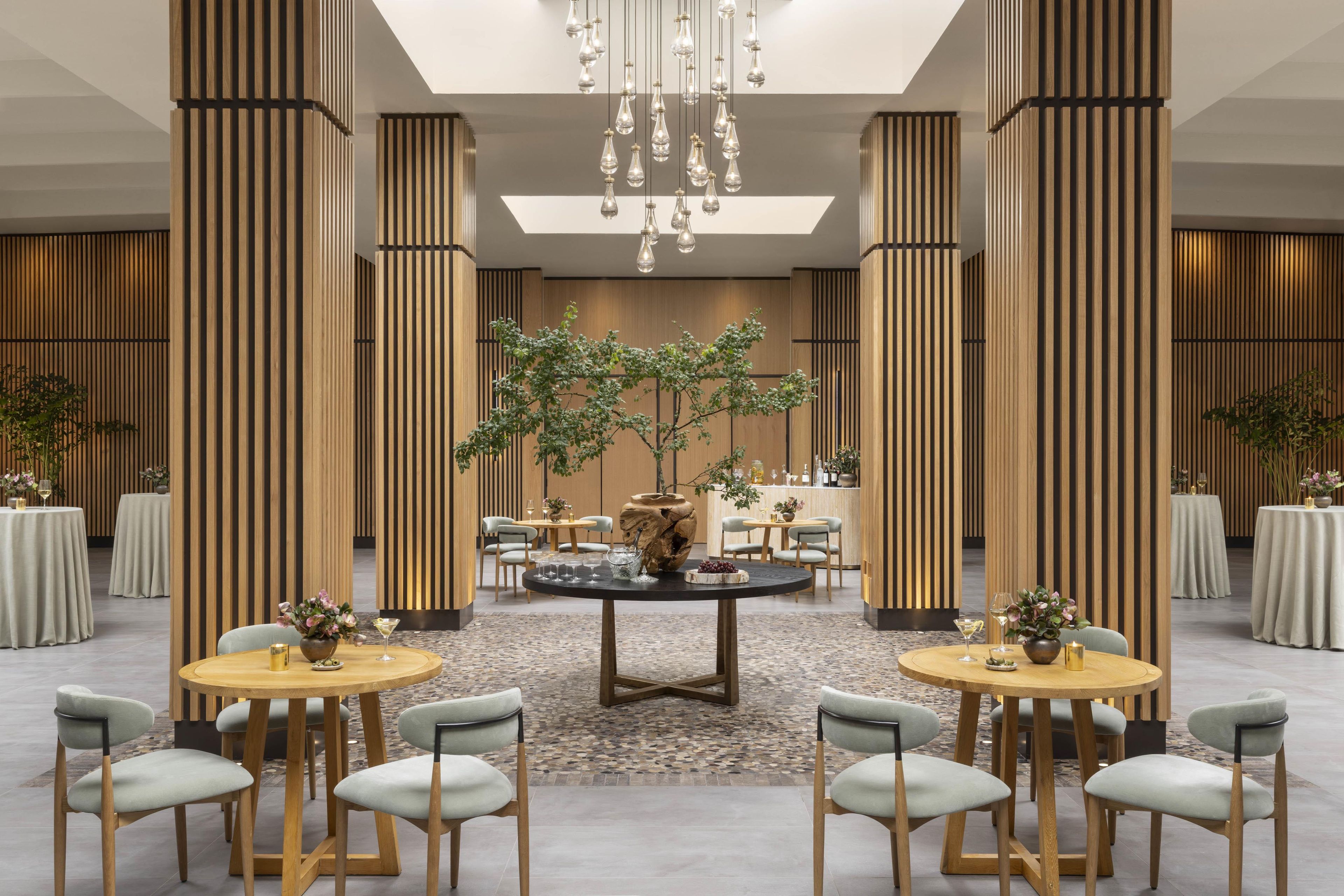 A spacious hotel lobby with high ceilings, a central round table surrounded by chairs, and vertical wooden panels.