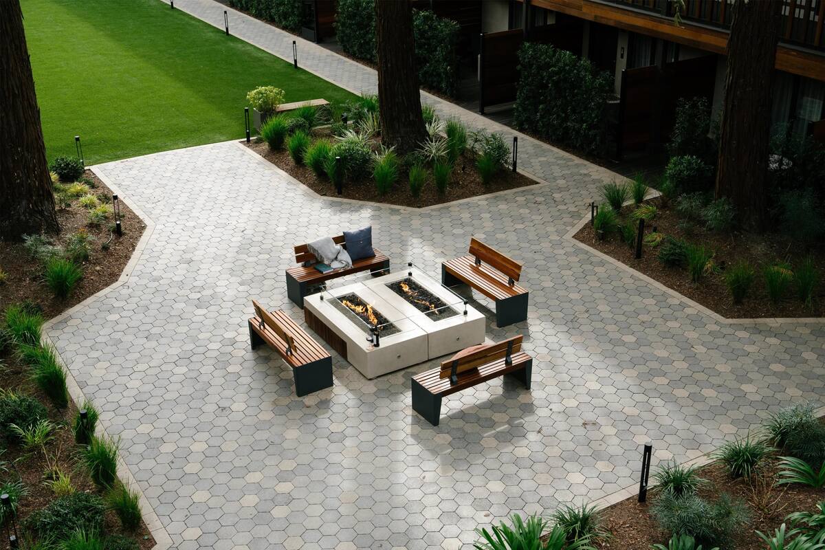 Eine sechseckige Terrasse mit Fliesen, die ein zentrales Feuerbecken hat, umgeben von Holzbanken und Pflanzgefäßen.