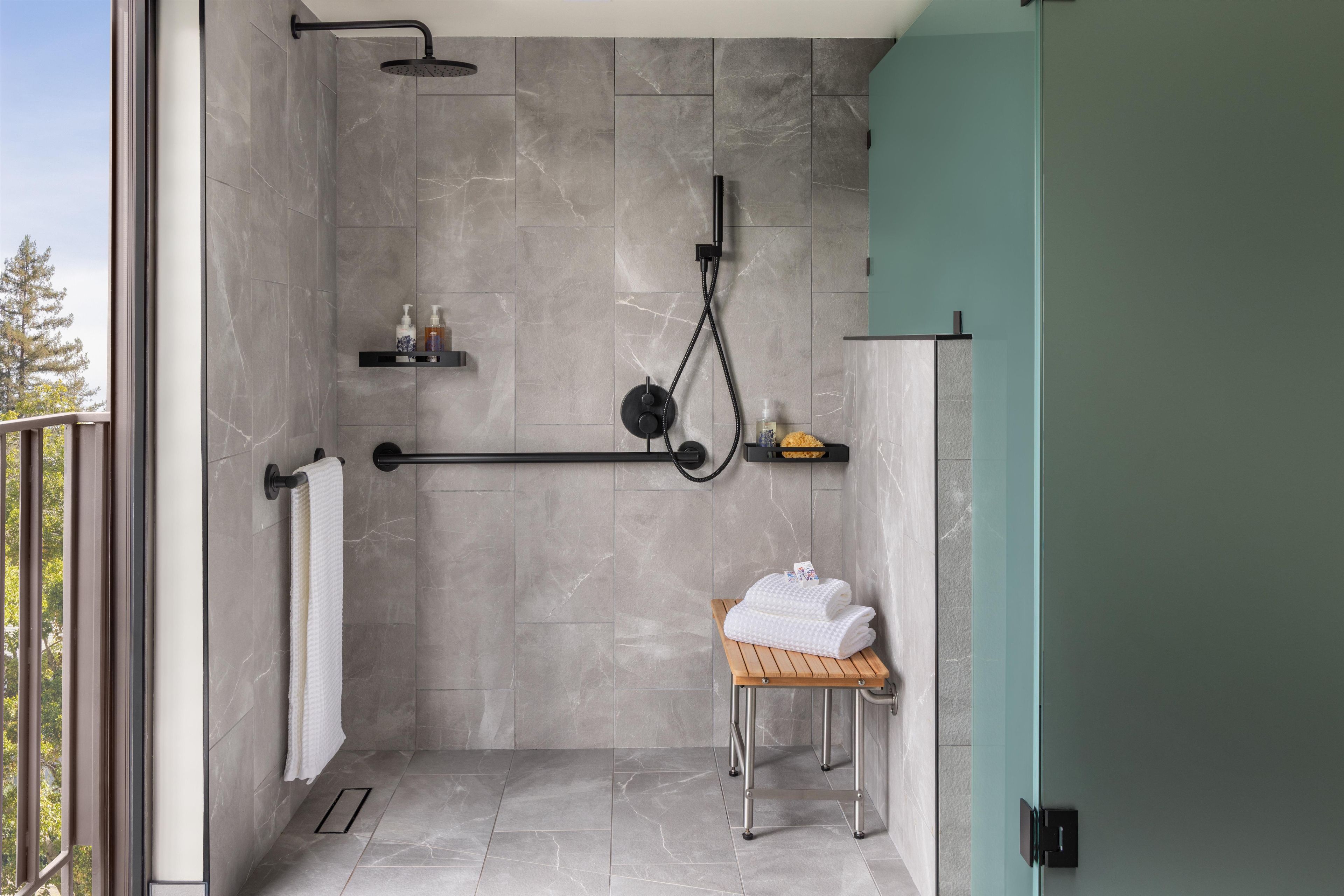 A modern shower with a rain showerhead, a handheld showerhead, and a handheld massager. There is a towel rack with a towel and a small wooden stool with towels.