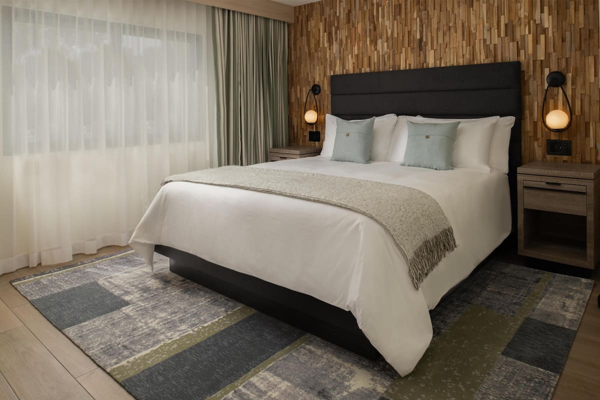 Ein modernes Hotelzimmer mit einem großen Bett, zwei Nachttischen, einer Wandlampe und einem Fenster mit transparenten Vorhängen.