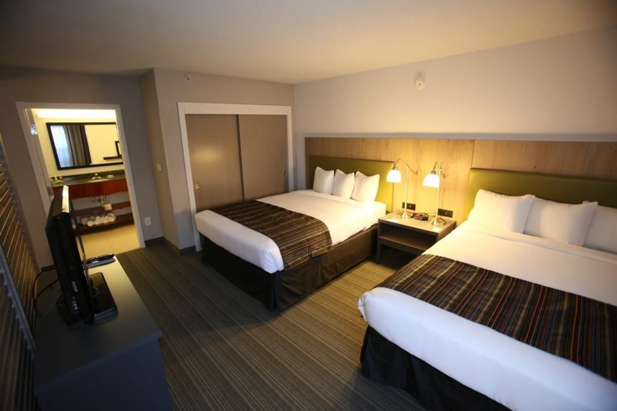 Habitación de hotel con dos camas, un escritorio, un espejo y un baño visible a través de una puerta abierta.