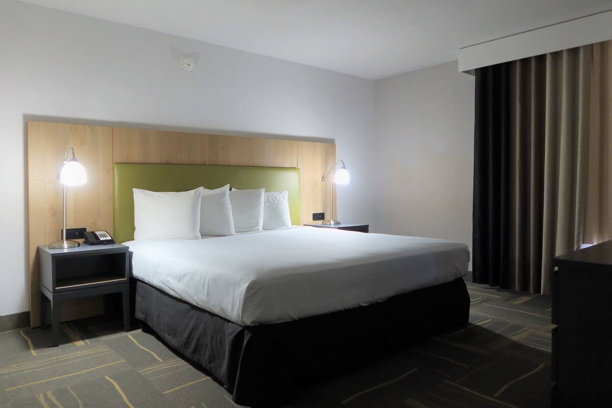 Une chambre d’hôtel avec un grand lit, deux tables de chevet avec des lampes, une tête de lit en bois et un rideau foncé à la fenêtre.