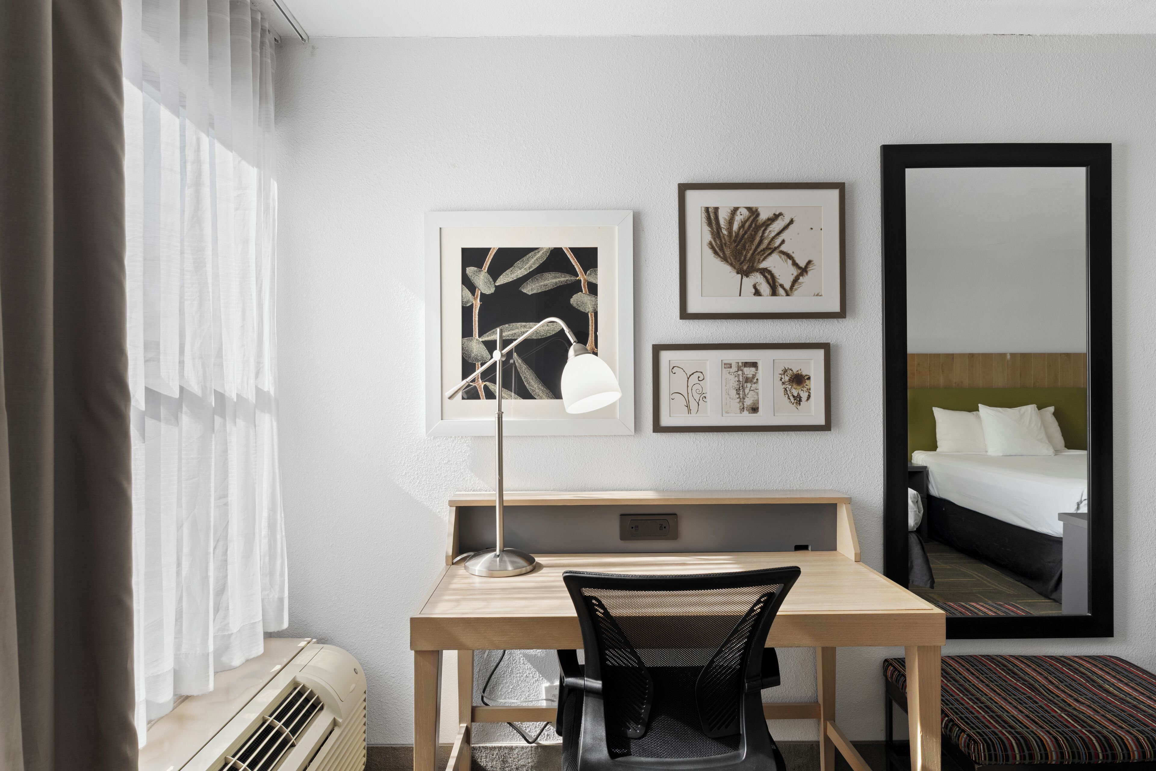 Une chambre d’hôtel équipée d’un bureau, d’une chaise et d’un grand miroir. Les murs sont ornés d’œuvres d’art encadrées, et il y a une fenêtre avec des rideaux.