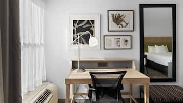 Habitación de hotel con escritorio, silla y un espejo grande. En la pared hay obras de arte enmarcadas y una ventana con cortinas.