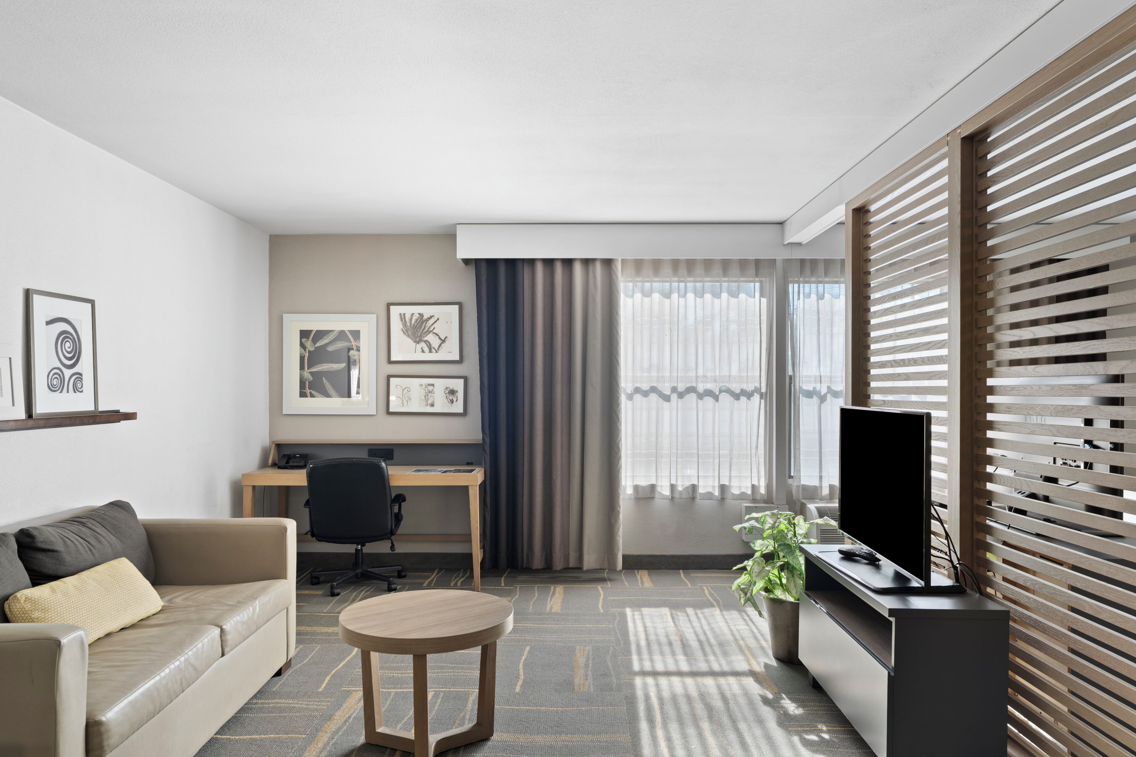 A modern hotel room with a sofa, a round coffee table, a desk with a chair, a television, and artwork on the walls.
