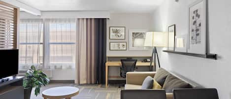 A hotel room with a TV, a dining table, a sofa, a desk with a chair, and a lamp.