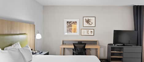A hotel room with a bed, desk, chair, TV, and artwork on the wall.