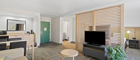 A modern hotel room with a kitchenette, a flat-screen TV, a seating area with a sofa and a small round table, and a large potted plant.