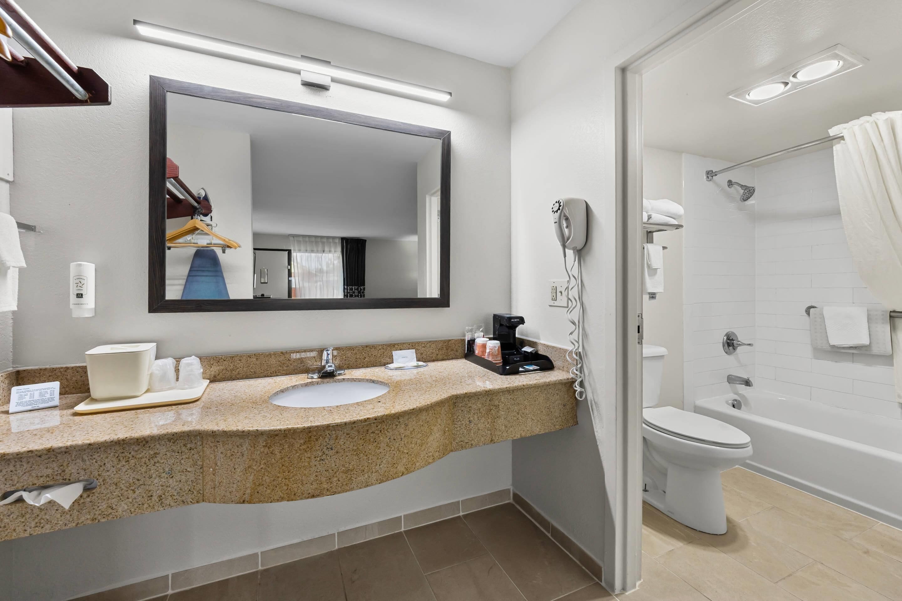 A modern bathroom with a granite countertop, a sink, a mirror, a hairdryer, and a shower area.