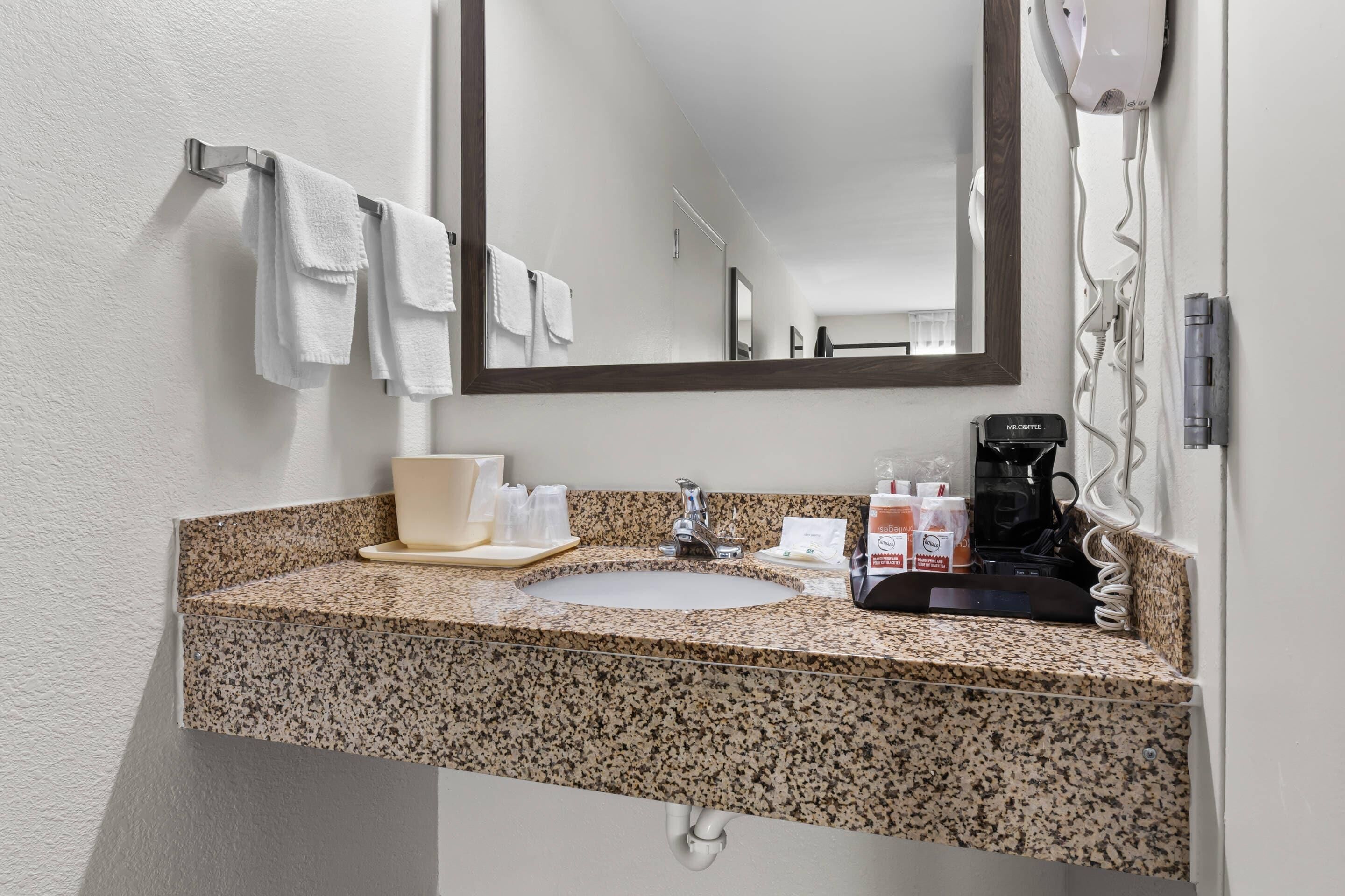A bathroom with a granite countertop, a sink, a mirror, a hairdryer, and a coffee maker.
