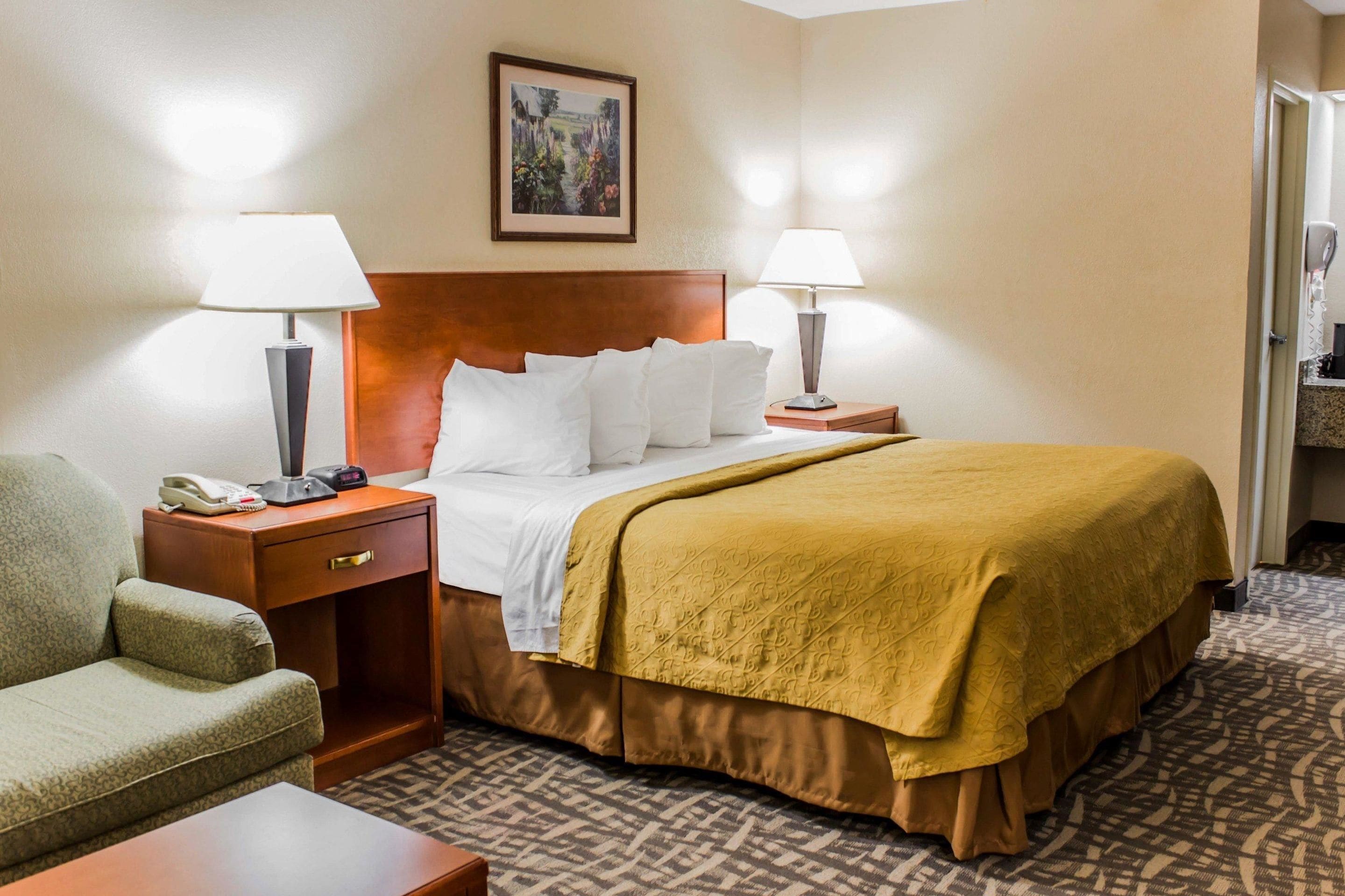 A hotel room with a bed, two bedside lamps, a nightstand with a phone, a chair, and a painting on the wall.