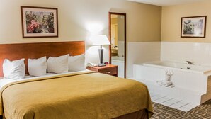 A hotel room with a large bed, a bedside table with a lamp, a bathroom with a bathtub, and two framed pictures on the walls.