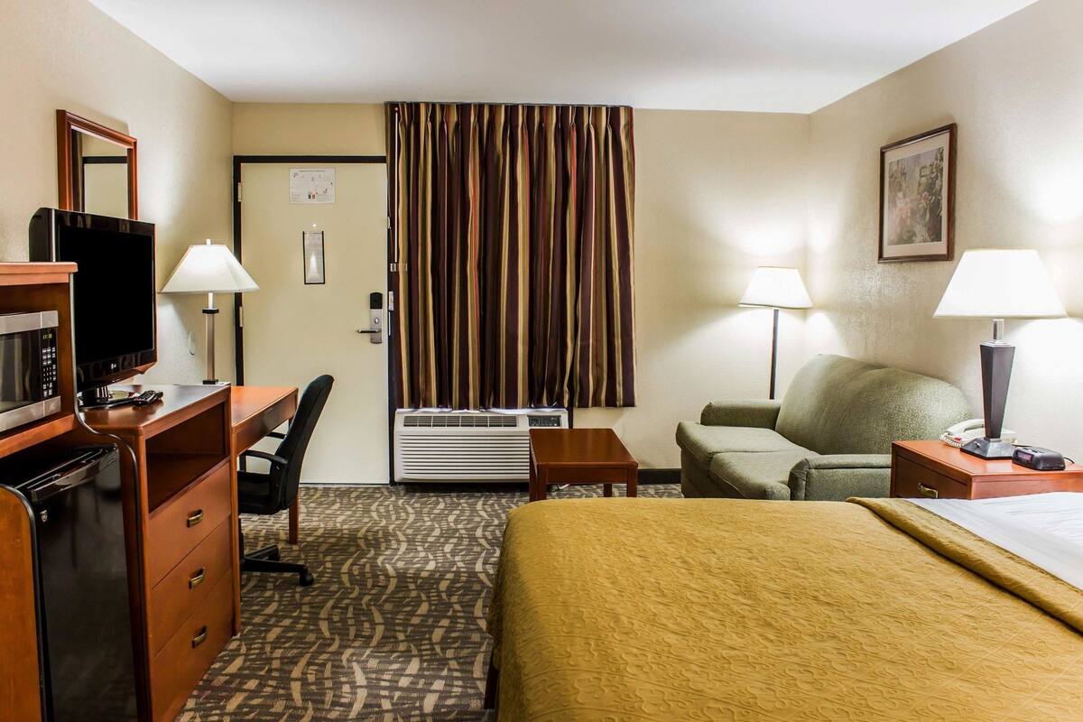 A hotel room with a bed, desk, chair, television, air conditioning unit, lamps, and a painting on the wall.