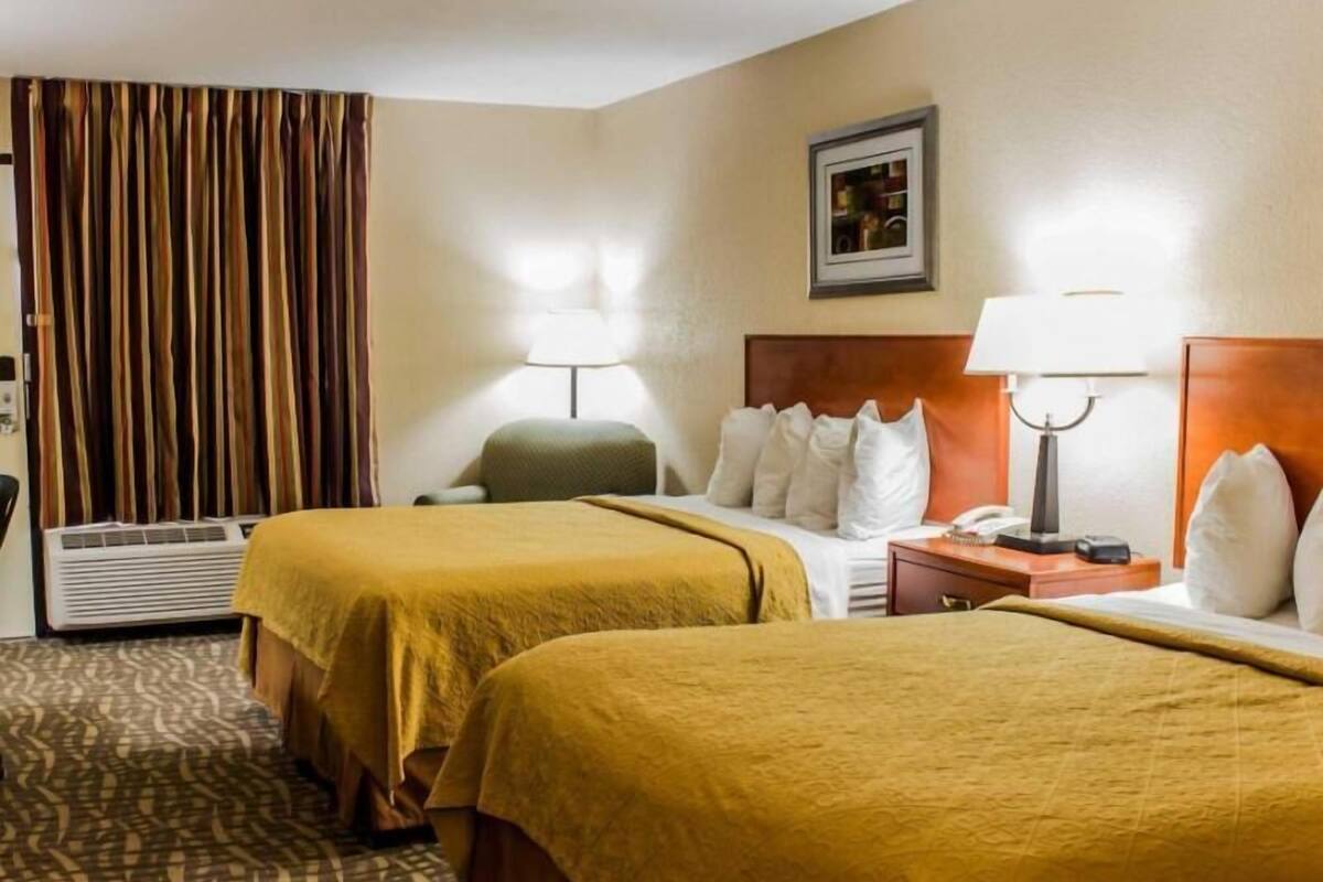 A hotel room with two beds, a desk, a chair, a lamp, a heater, and a picture on the wall.
