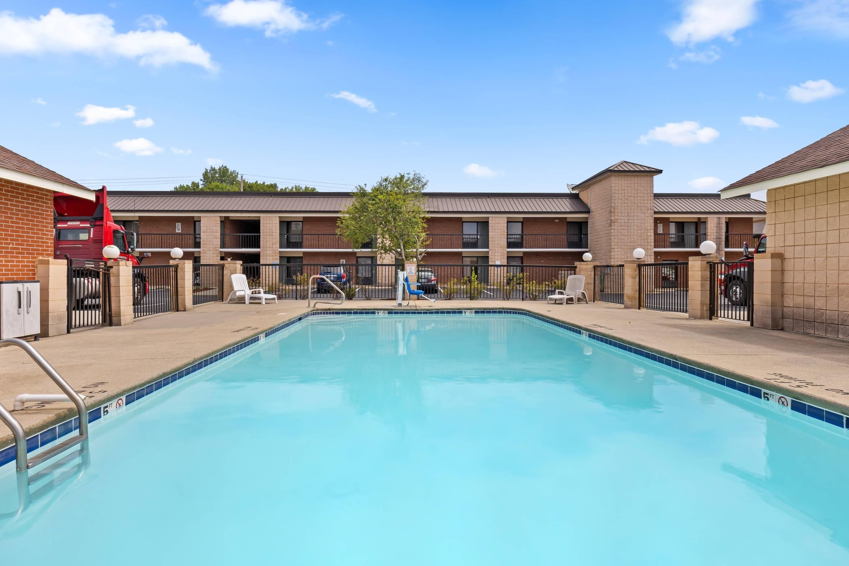 A rectangular swimming pool with a clear blue water surface, surrounded by a low brick wall, and a building with a flat roof in the background.