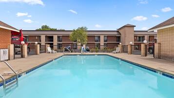 A rectangular swimming pool with a clear blue water surface, surrounded by a low brick wall, and a building with a flat roof in the background.
