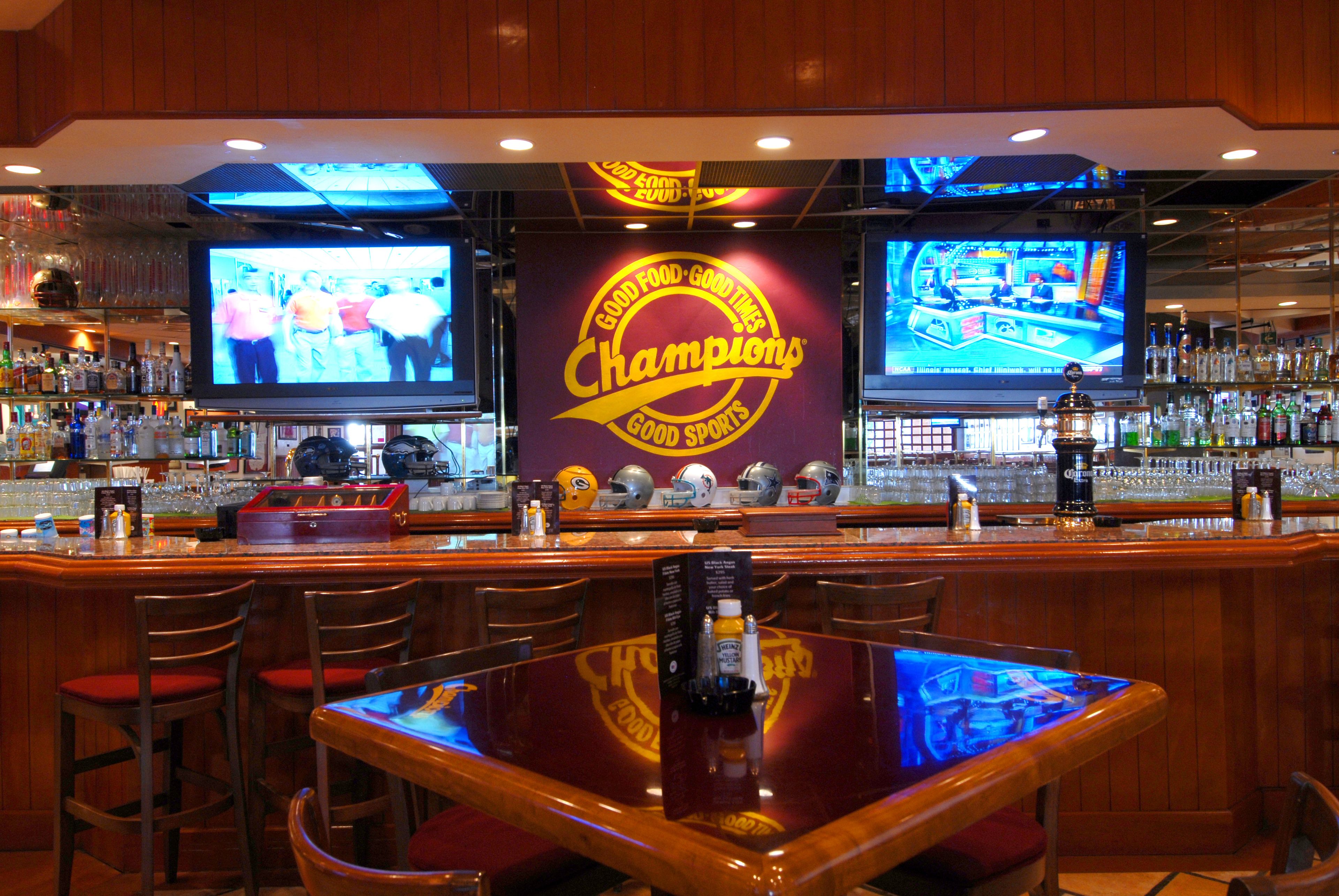 Uma área de bar com várias televisões exibindo eventos esportivos, um balcão com diversos tipos de bebidas e um grande logotipo na parede.