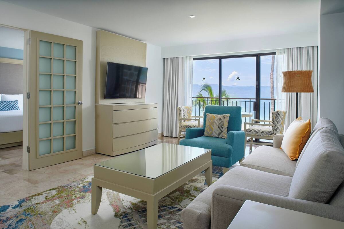 A hotel room with a sofa, a television, a coffee table, a balcony with a view, and a bed with a sliding door.