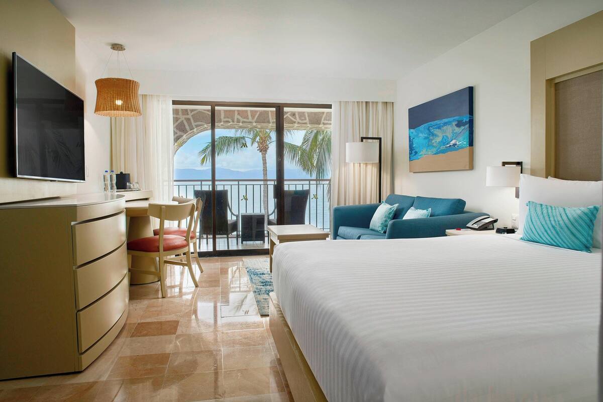 A hotel room with a bed, a desk with a chair, a TV, a balcony with a view of palm trees and the ocean, and a painting on the wall.