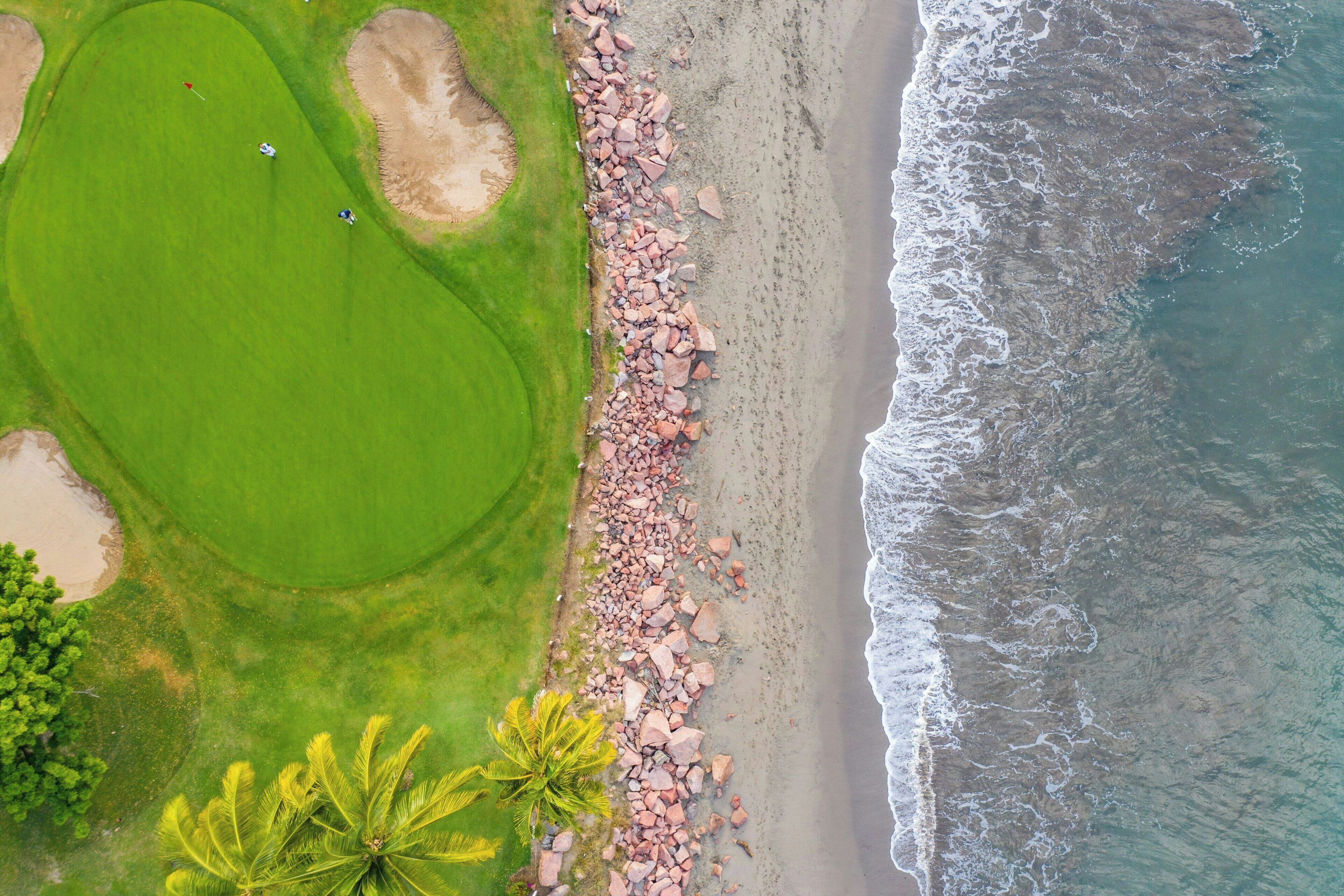 An aerial view of a coastal golf course with a sandy beach and rocky shoreline.