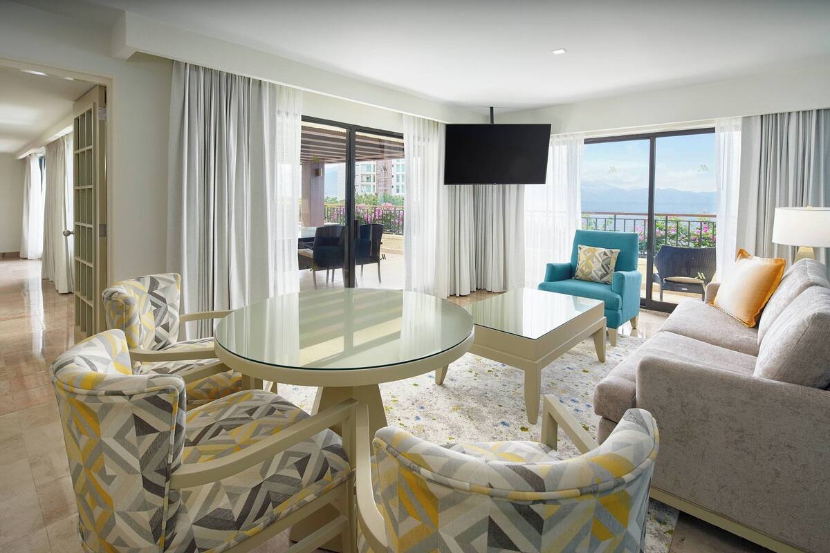 A hotel room with a glass dining table, patterned chairs, a sofa, a TV, and a balcony with outdoor seating.