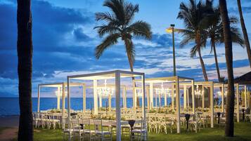 An outdoor dining area with white tables and chairs, surrounded by palm trees, overlooking the sea.