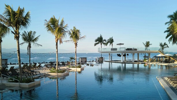 Ein Poolbereich mit Liegestühlen, Palmen und Blick aufs Meer.