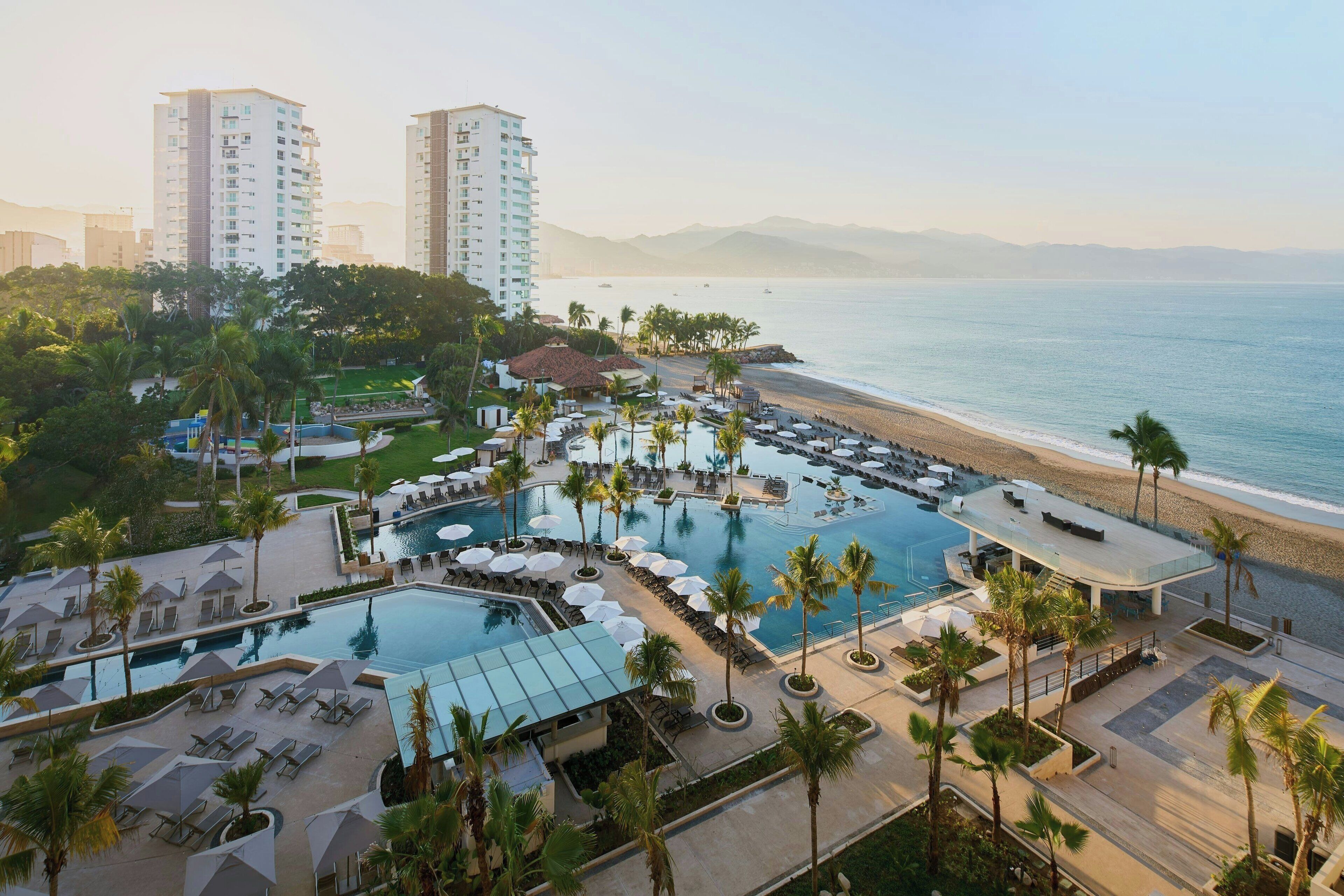 A resort with a large pool, beachside lounge areas, and high-rise buildings in the background.