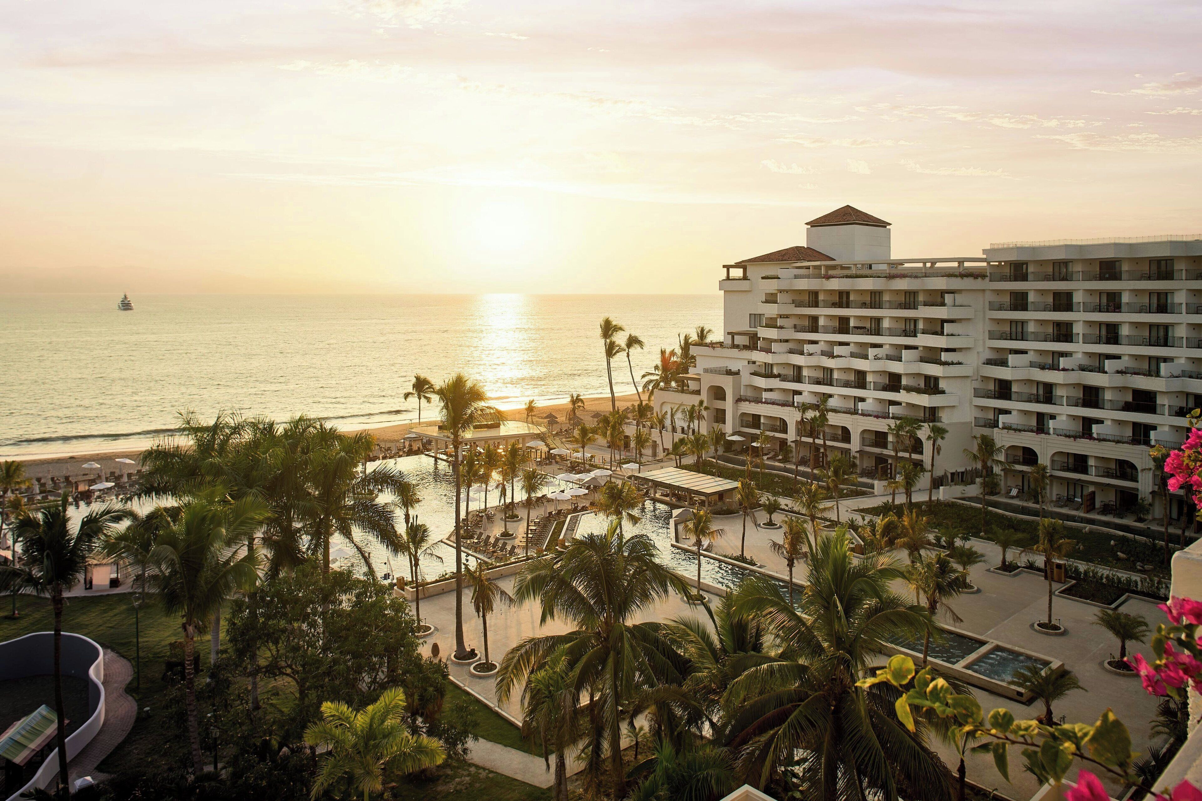 A resort with a pool area, palm trees, and a view of the ocean at sunset.