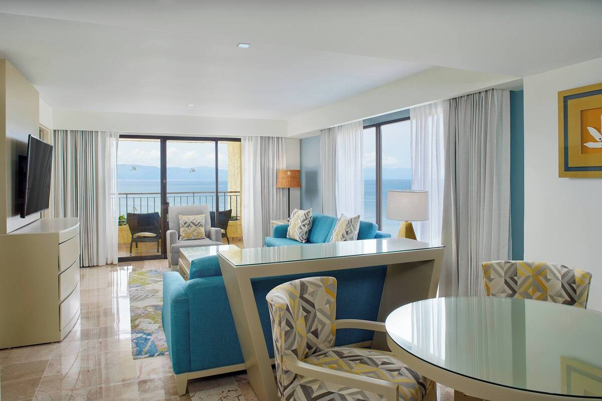 A modern hotel room with a balcony, a TV, a sofa, a dining table, and a chair.