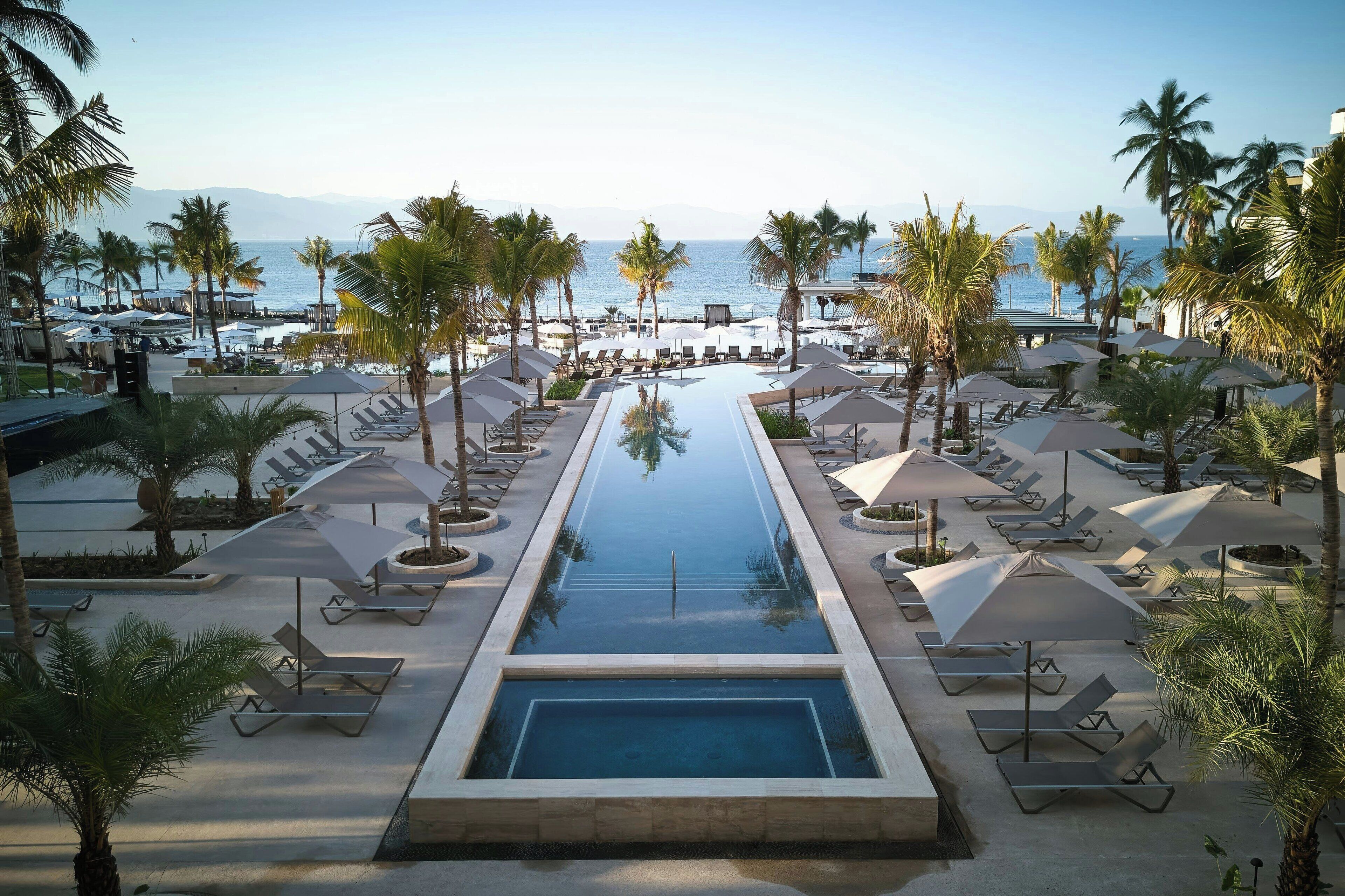 A pool area with lounge chairs and umbrellas, surrounded by palm trees, overlooking a beach and ocean.