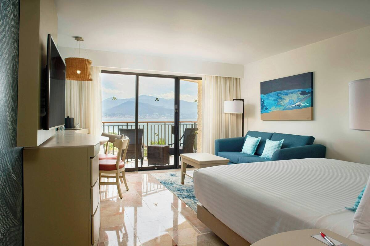 A hotel room with a bed, a desk, a chair, a sofa, a TV, and a balcony with a view of mountains.